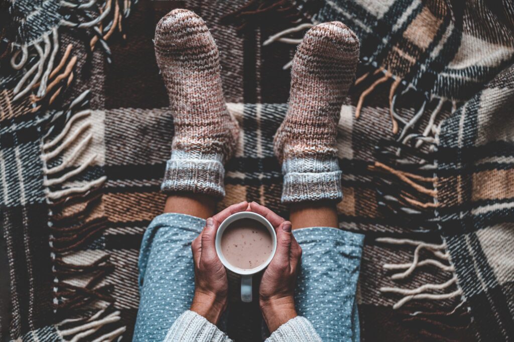 Cozy woman in knitted winter warm socks and in pajamas holding a cup of hot cocoa during resting on checkered plaid blanket at home in winter time. Cozy time and winter drinks. Top view